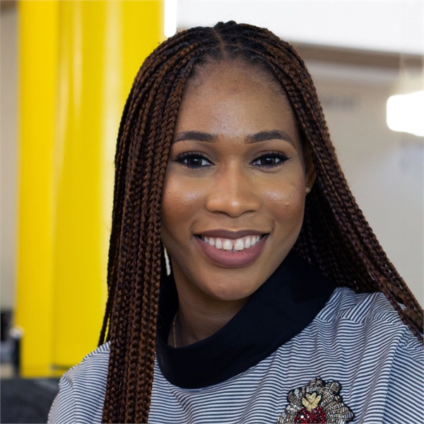 Chisom, a young black woman with braids smiling at the camera. She's wearing a high collared top with a gold, silver and ruby red crest on the breast.