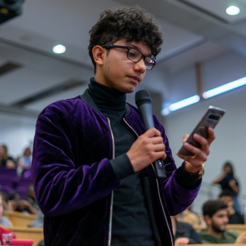 Student standing, talking to a microphone while reading something from his phone.