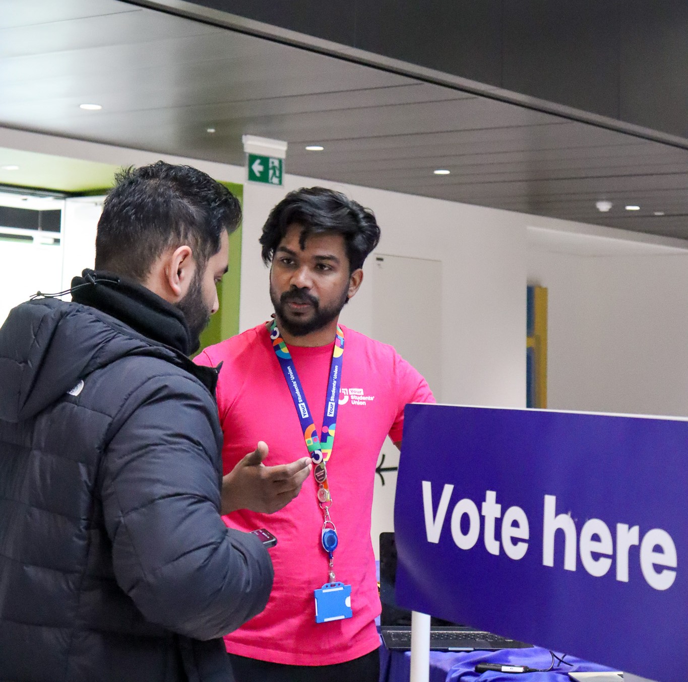Two people having a conversation with a sign that reads: "vote here".