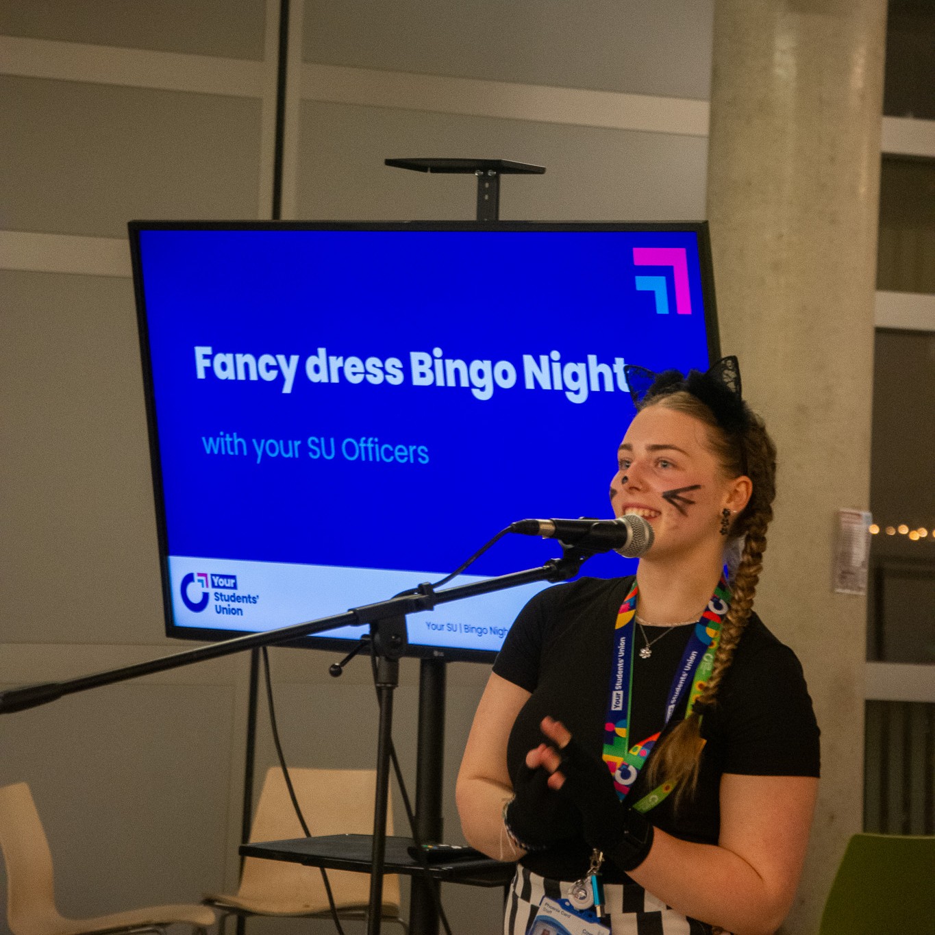 Person speaking into a microphone with a screen in the background reading 'Fancy dress bingo night with your SU Officers.'